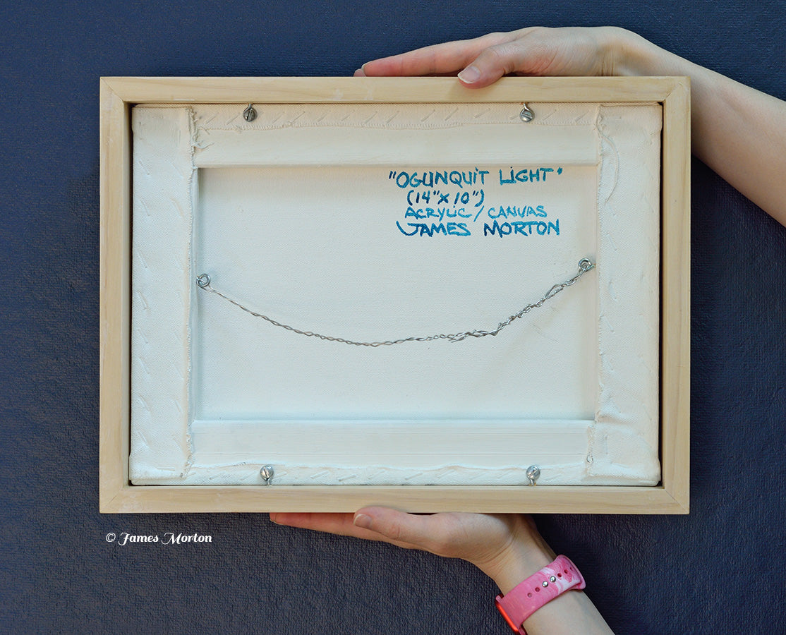 Back of framed original acrylic painting held for scale showing the handwritten title "Ogunquit Light," artist signature, 14 × 10 canvas, hanging wire, and natural wood frame.