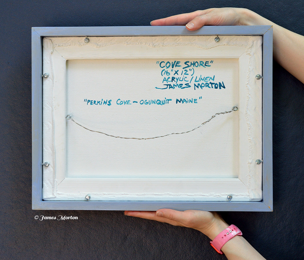 Back of framed original acrylic painting on linen titled Cove Shore, depicting Perkins Cove in Ogunquit, Maine. Hand signed, titled, labeled by the artist, wired and ready to hang, shown held for scale.