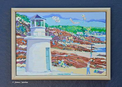 Framed original Maine lighthouse painting by James Morton depicting Lobster Point Lighthouse along Ogunquit's Marginal Way with a rocky shoreline, blue ocean, beachgoers, and colorful kites.