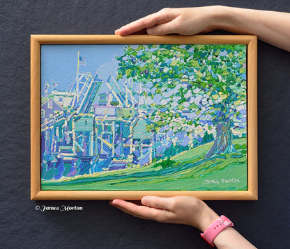 Original Ogunquit Maine acrylic painting, 14 x 10 inches, shown held for scale. Framed Perkins Cove landscape featuring the historic drawbridge, large shade tree, blue sky, and coastal architecture by James Morton.