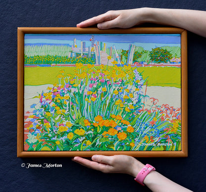 Framed original acrylic painting of a colorful coastal garden in Wellfleet, Massachusetts, shown held for scale. The scene features yellow, orange, pink, and blue blossoms, green grass, and a small wooden boat dock in the background.