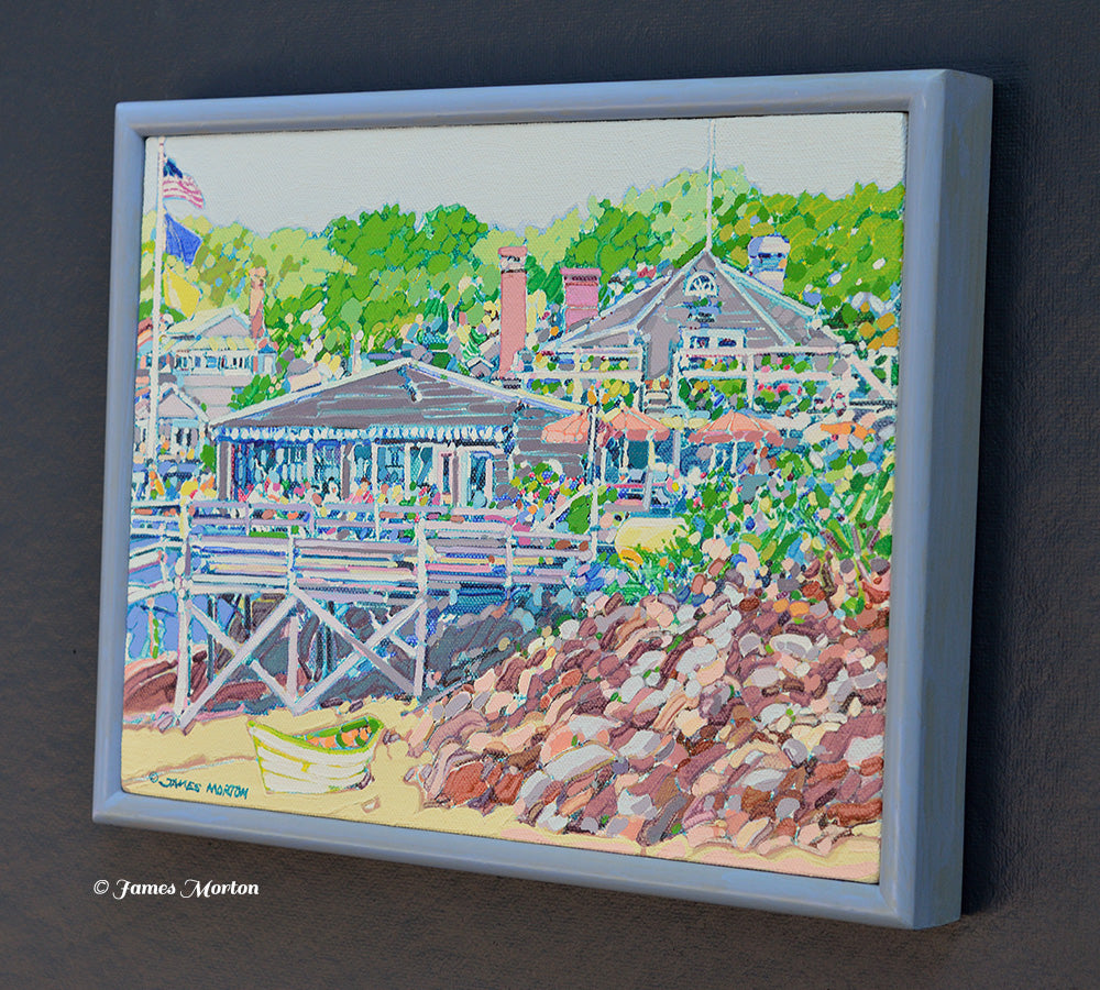 Right side angle view of a framed original New England coastal painting depicting Barnacle Billy's restaurant, rocky shoreline, white rowboat, beach, and lush trees at Perkins Cove in Ogunquit, Maine.