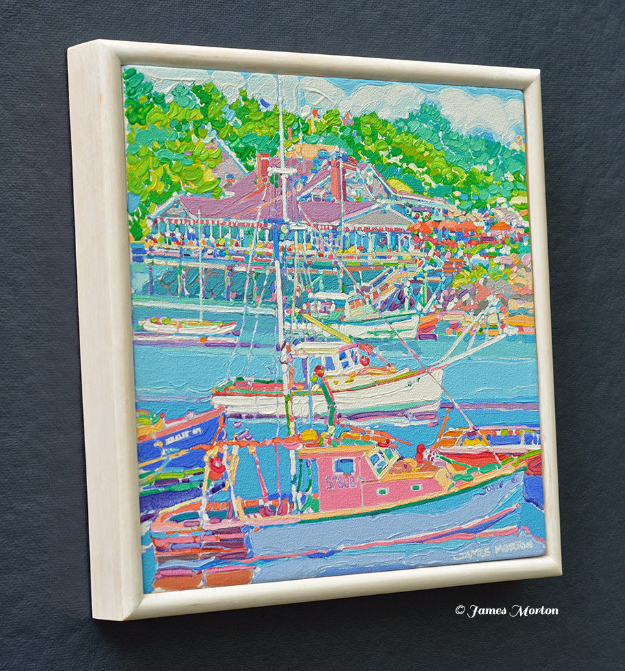 A framed original painting of Ogunquit Maine with fishing boats, green trees, and harbor side restaurant. The frame is white stained wood, and painted signature James Morton appears at the bottom right.