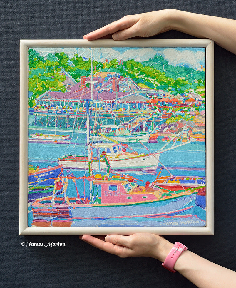 Coastal Maine Painting of Perkins Cove harbor scene with boats, restaurant and green trees, 14x14 size shown held by hands for scale The artwork has a white stained wood frame, and the signature 'James Morton' is visible in the lower right corner of the painting.