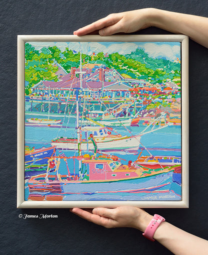 Coastal Maine Painting of Perkins Cove harbor scene with boats, restaurant and green trees, 14x14 size shown held by hands for scale The artwork has a white stained wood frame, and the signature 'James Morton' is visible in the lower right corner of the painting.
