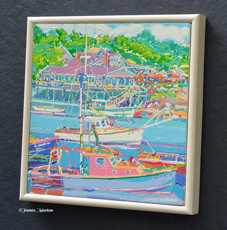 Right angle view framed Ogunquit Maine painting of a Perkins Cove harbor scene with boats, outdoor restaurant, green trees, The cream-colored wood frame is visible, and a small artist signature 'James Morton' appears in the lower right corner of the artwork.