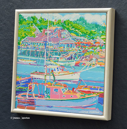 Right angle view framed Ogunquit Maine painting of a Perkins Cove harbor scene with boats, outdoor restaurant, green trees, The cream-colored wood frame is visible, and a small artist signature 'James Morton' appears in the lower right corner of the artwork.
