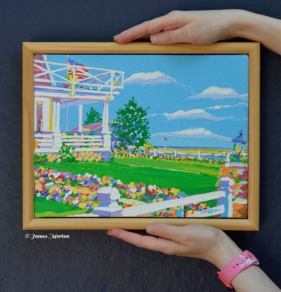 Hand-held view of a framed 12 x 9 original acrylic painting of Ogunquit, Maine for size reference. The artwork features a flower-lined pathway leading to a classic New England inn with wrap-around porch, green lawn, coastal trees, and blue sky.