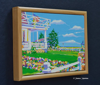 Left side angle of a framed original Maine coastal painting featuring a white Colonial inn, colorful flower garden, wrap-around porch, trees, bright blue sky, and hints of the Ogunquit coastline. Light pine wood frame with artist signature visible.
