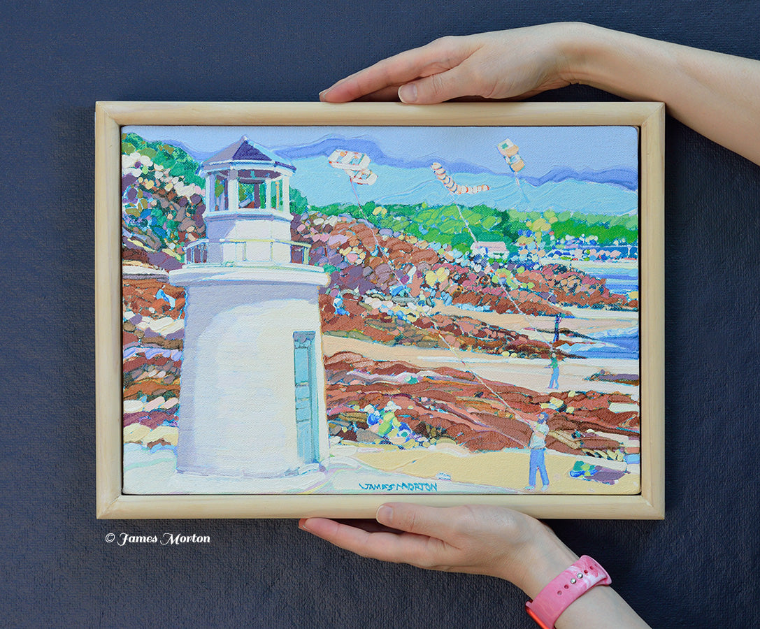 Framed 14 × 10 original acrylic painting held for scale featuring Lobster Point Lighthouse along Ogunquit's Marginal Way with a rocky shoreline, beach, blue sky, and colorful kites.